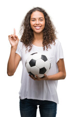 Wall Mural - Young hispanic woman holding soccer football ball surprised with an idea or question pointing finger with happy face, number one