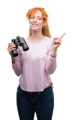 Poster - Young redhead woman looking through binoculars very happy pointing with hand and finger to the side