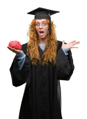 Poster - Young redhead student woman wearing graduated uniform holding brain very happy and excited, winner expression celebrating victory screaming with big smile and raised hands