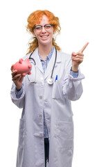 Poster - Young redhead doctor woman holding piggy bank very happy pointing with hand and finger to the side