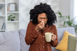 Young woman feeling unwell with cold or flu, sneezing into tissue, holding mug, at home on cozy sofa with cushions