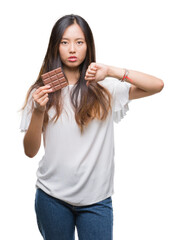 Wall Mural - Young asian woman eating chocolate bar over isolated background with angry face, negative sign showing dislike with thumbs down, rejection concept