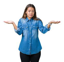 Wall Mural - Young beautiful brunette woman wearing blue denim shirt over isolated background clueless and confused expression with arms and hands raised. Doubt concept.