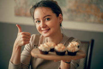 Wall Mural - A young woman shows a cool sign with cupcakes in her hand. cream decoration. The bakery chef bakes cakes in the kitchen