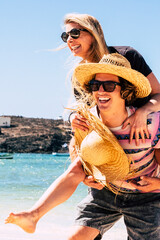 Wall Mural - Young man laughing with girlfriend in piggyback - summer holiday vacation people with beach and blue sea water in background - youthful couple have fun together outdoor