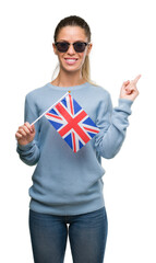 Wall Mural - Beautiful young woman holding UK flag very happy pointing with hand and finger to the side