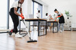 © sofiko14 - Cropped view of hands of African man holding vacuum cleaner. Team of young multicultural cleaners vacuums floor, wipes tables and gadgets, shelves, washes windows in spacious, bright, modern office.