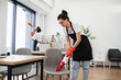 © sofiko14 - Multiethnic young female cleaner vacuuming chair with portable cordless vacuum cleaner. Happy smiling multicultural employees of company cleaning in bright, spacious modern office.