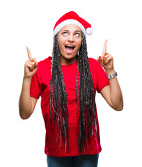 Canvas Print - Young braided hair african american girl wearing christmas hat over isolated background amazed and surprised looking up and pointing with fingers and raised arms.