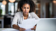 © MP Studio - Focused woman with curly hair, wearing a white shirt, reading a document in a bright, home office setting, with a laptop open in front of her.