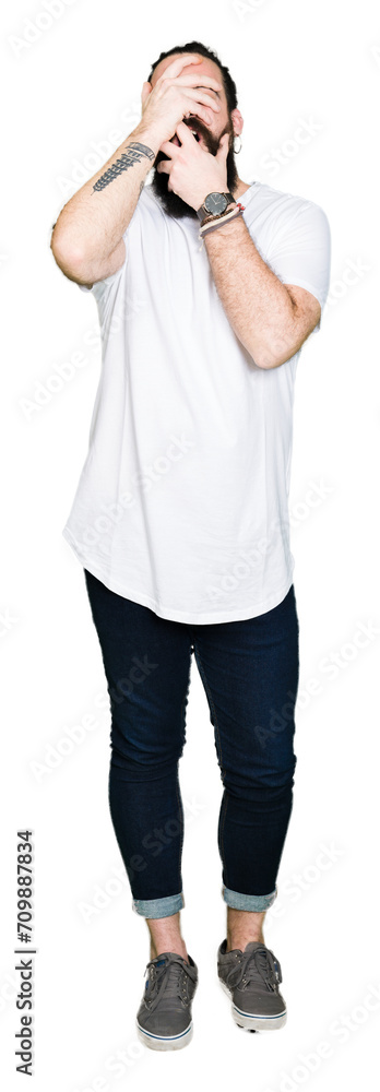 Young hipster man with long hair and beard wearing casual white t-shirt Covering eyes and mouth with hands, surprised and shocked. Hiding emotion