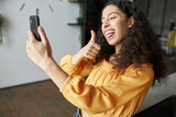 Fototapeta  - Side view of funny black girl of 20s recording short video or taking selfie on smartphone grimacing, making faces, sticking tongue and showing thumb up, having fun being in good mood