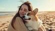 A girl in a beige sweater hugs a corgi dog sitting on the sand by the sea