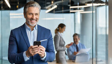 Smiling Older Business Man Executive Standing In Office Using Mobile Phone. Happy Mid Aged Professional Manager Holding Cell Working On Smartphone Checking Financial Transactions On Cellphone Tech