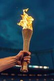 Fototapeta  - Hand holding flaming Olympic torch with Olympic stadium in the background