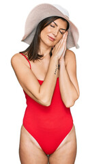 Wall Mural - Beautiful brunette woman wearing swimsuit and summer hat sleeping tired dreaming and posing with hands together while smiling with closed eyes.