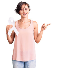 Wall Mural - Beautiful young woman with short hair holding cancer awareness white ribbon smiling happy pointing with hand and finger to the side