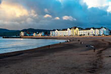 A Beach In Golden Panoramic Landscape View. Orange Dusk Sunlight Is Cast Across The Beach And Ornate White Buildings. People Walking On The Beach.