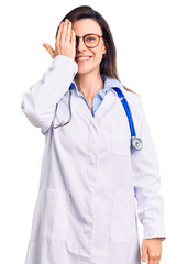 Wall Mural - Young beautiful woman wearing doctor stethoscope and glasses covering one eye with hand, confident smile on face and surprise emotion.