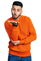 Wall Mural - Young hispanic man with beard wearing casual orange sweater looking at the camera blowing a kiss with hand on air being lovely and sexy. love expression.