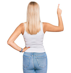 Canvas Print - Young blonde girl wearing casual style with sleeveless shirt posing backwards pointing ahead with finger hand