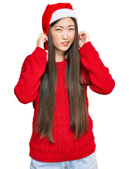 Sticker - Young chinese woman wearing christmas hat covering ears with fingers with annoyed expression for the noise of loud music. deaf concept.