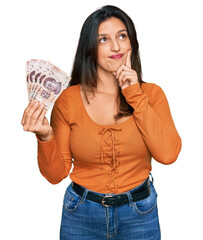 Canvas Print - Beautiful hispanic woman holding mexican pesos serious face thinking about question with hand on chin, thoughtful about confusing idea