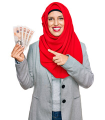 Canvas Print - Beautiful hispanic woman wearing islamic hijab holding 10 pounds banknotes smiling happy pointing with hand and finger