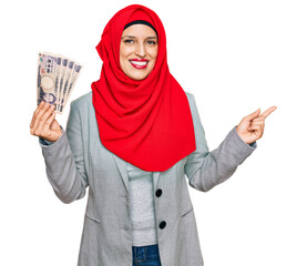 Wall Mural - Beautiful hispanic woman wearing islamic hijab holding japanese yen banknotes smiling happy pointing with hand and finger to the side