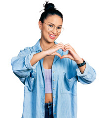 Wall Mural - Beautiful hispanic woman wearing casual denim jacket and glasses smiling in love doing heart symbol shape with hands. romantic concept.