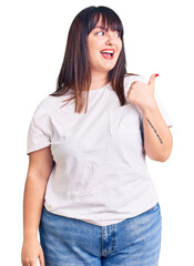 Sticker - Young plus size woman wearing casual clothes smiling with happy face looking and pointing to the side with thumb up.