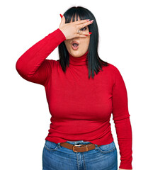 Sticker - Young hispanic woman wearing casual clothes peeking in shock covering face and eyes with hand, looking through fingers with embarrassed expression.