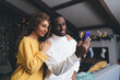Loving interracial couple engaged with mobile phone; woman in sweater rests her head on man in white turtleneck, both content in festive home setting look at smartphone screen content in social media