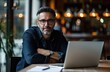© Lars - A smiling middle-aged man, donned in a casual jeans shirt and glasses, exudes confidence while sitting behind a laptop, making direct eye contact with the camera.