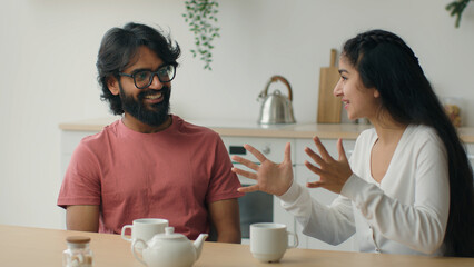 Happy multiracial couple drink coffee tea in home kitchen friendly chatter Arabian man guy listen funny story carefree conversation Indian woman girl talk storytelling romantic date friends friendship