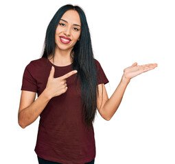 Wall Mural - Young hispanic girl wearing casual t shirt amazed and smiling to the camera while presenting with hand and pointing with finger.