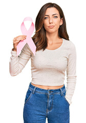 Wall Mural - Young brunette woman holding pink cancer ribbon thinking attitude and sober expression looking self confident