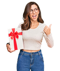 Sticker - Young brunette woman holding gift pointing thumb up to the side smiling happy with open mouth