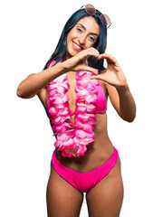 Wall Mural - Beautiful hispanic woman wearing bikini and hawaiian lei smiling in love doing heart symbol shape with hands. romantic concept.