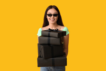 Sticker - Beautiful young Asian woman with gift boxes on yellow background. Black Friday sale