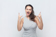 Woman showing rock and roll gesture, heavy metal sign, enjoying favorite music on party, sticking tongue, exclaims from joy, wearing striped dress. Indoor studio shot isolated on gray background.