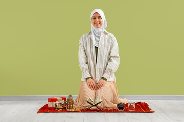 Wall Mural - Young Muslim woman praying on mat near green wall. Ramadan celebration