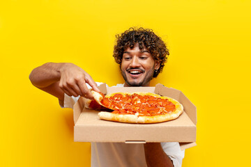 Wall Mural - young hungry indian guy holding a box of delicious pizza and taking a piece on a yellow isolated background, curly happy man eating fastfoot and smiling