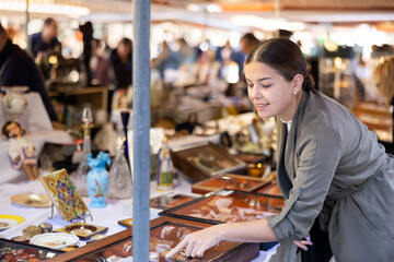 Satisfied interested young woman choosing interesting antique things at traditional flea market