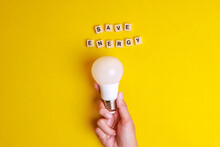 Save Energy Words Written On Wooden Blocks With Hand Holding White Light Bulb On Yellow Background. Concept Of Saving Energy By Using LED Lightbulb