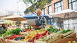 © Gorodenkoff - Portrait of a Middle Aged Street Vendor Working on Laptop Computer while Standing in a Farmers Market Stall with Fresh Natural Agricultural Products. Businessman Contacting Suppliers Online