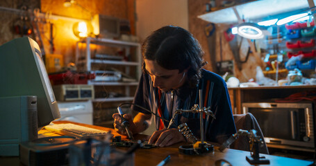 Leinwandbilder - Portrait Of Caucasian Male Engineer Using Soldering Iron On Circuit Board In Front Of Old Desktop Computer In Nineties Garage. Engineering Student Working On Project For College At Home In the Evening