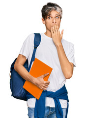 Wall Mural - Young hispanic man wearing student backpack and holding books covering mouth with hand, shocked and afraid for mistake. surprised expression