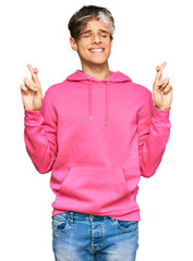 Poster - Young hispanic man wearing casual pink sweatshirt gesturing finger crossed smiling with hope and eyes closed. luck and superstitious concept.