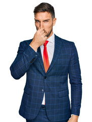 Poster - Handsome man with beard wearing business suit and tie smelling something stinky and disgusting, intolerable smell, holding breath with fingers on nose. bad smell